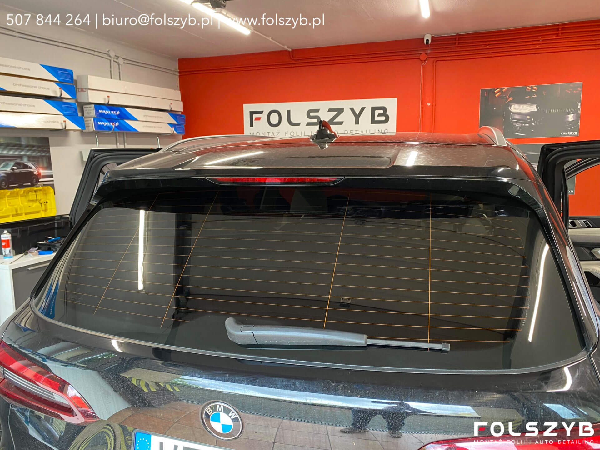 Tylna szyba BMW z folią, przez którą widać poziome ścieżki ogrzewania; hala FOLSZYB, na regale kartony z rolkami folii